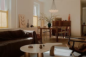Cozy living room with a sofa, coffee table, dining set, and decorative plants, lit by soft natural light from large windows.