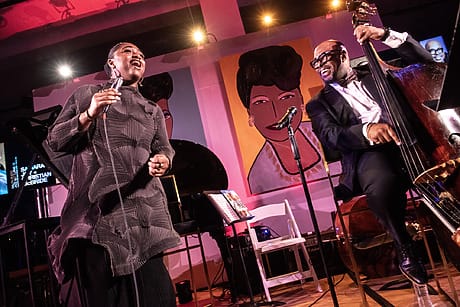 Musician sings while another plays double bass on stage, with bright portraits and lighting in the background.