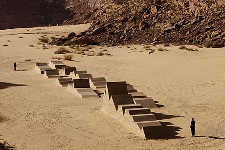 Concrete art installation in a desert landscape with two people standing at a distance on sand and rocky terrain.