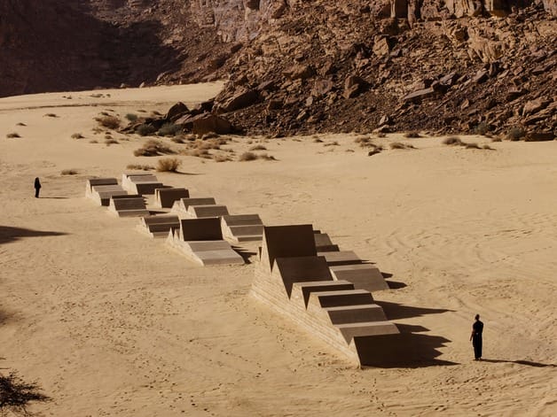 Concrete art installation in a desert landscape with two people standing at a distance on sand and rocky terrain.