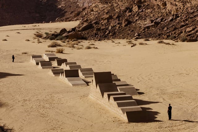 Concrete art installation in a desert landscape with two people standing at a distance on sand and rocky terrain.