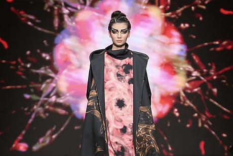 Model walking runway in colorful, artistic outfit with abstract background design during a fashion show.