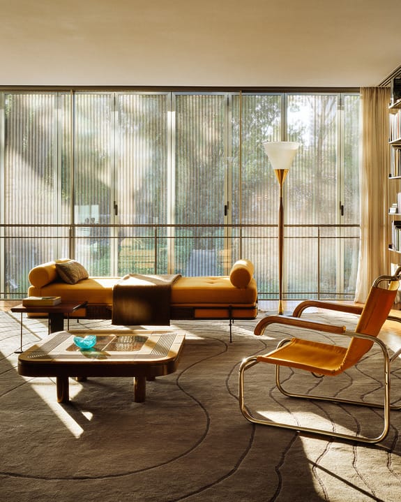 Modern living room with large windows, yellow chaise lounge, armchair, coffee table, and floor lamp in sunlight.
