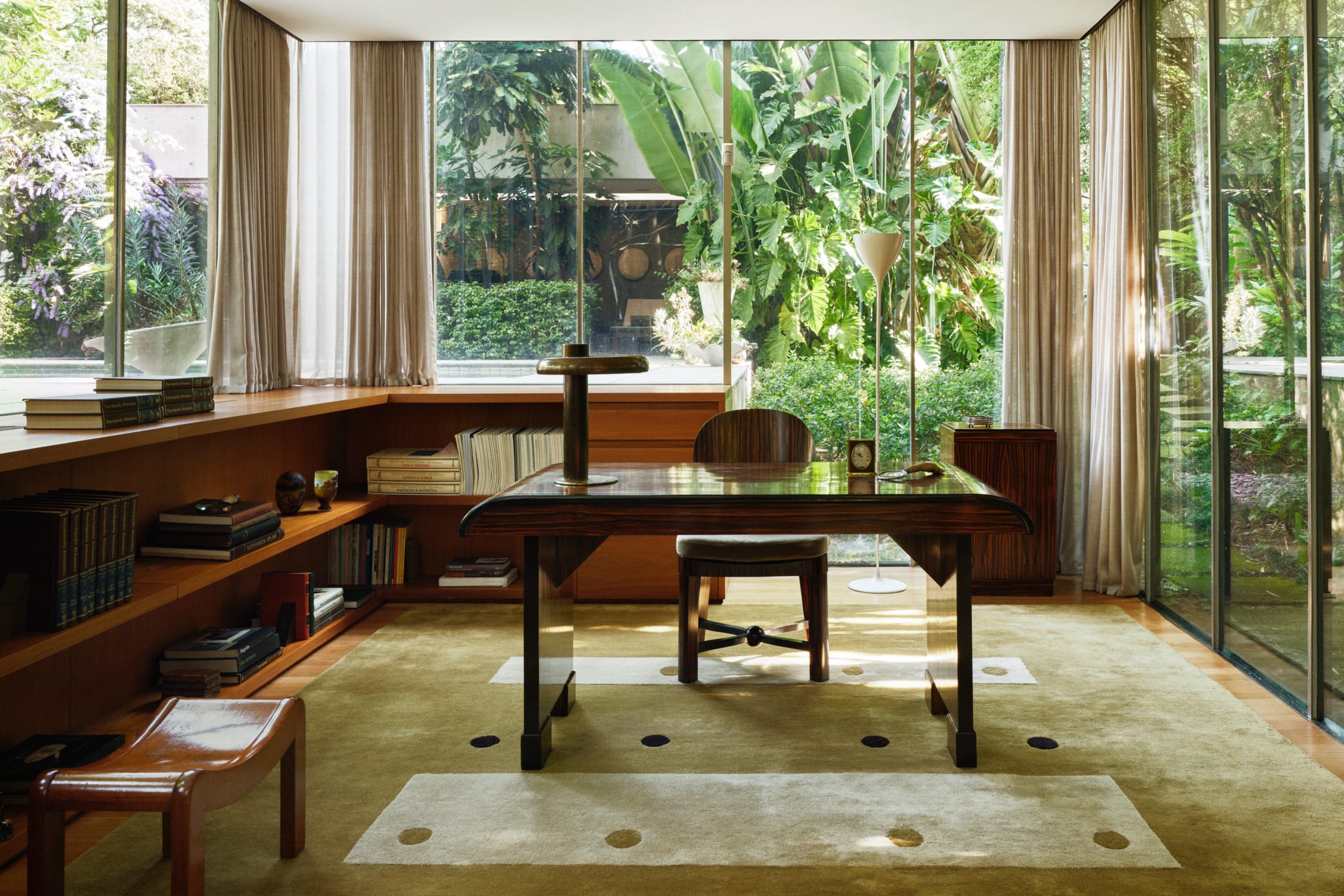 Mid-century modern office with large windows, surrounded by lush greenery, featuring a wooden desk and bookshelves.