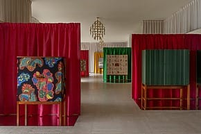 Art exhibition with colorful cabinets and vibrant curtains in a spacious room with chandeliers in the background.