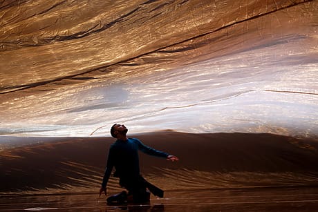 Person kneeling under a large, billowing golden fabric on a stage, looking upwards.