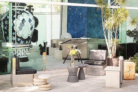 Outdoor patio with modern chairs, small tables, and a floral arrangement, in front of a glass wall showcasing contemporary art inside.