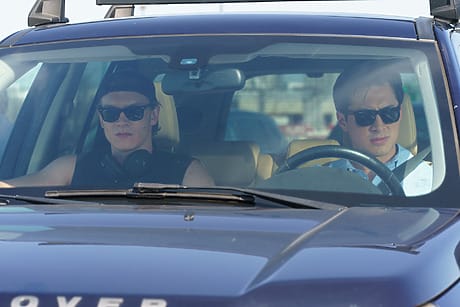 Two people wearing sunglasses inside a car, with one driving and the other in the passenger seat, both looking forward.