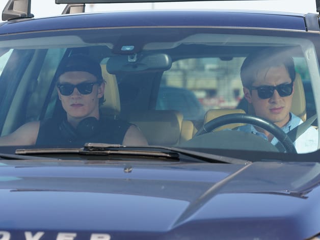 Two people wearing sunglasses inside a car, with one driving and the other in the passenger seat, both looking forward.