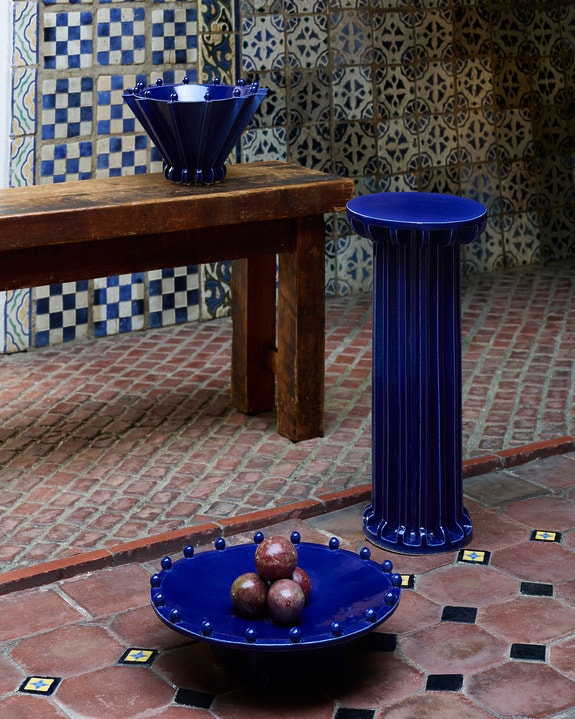 Blue ceramic bowl and pedestal on a red tiled floor with patterned walls in the background.
