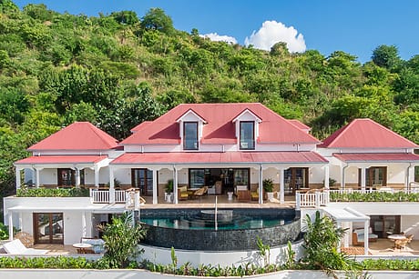 Large villa with red roof, infinity pool, and lush greenery on a hillside backdrop.