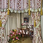 Decorated cozy room with floral drapes, wooden chair, desk, lamp, paintings, and vase of flowers, creating a vintage atmosphere.
