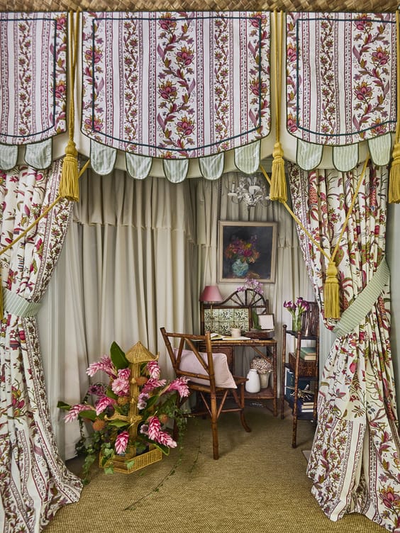 Decorated cozy room with floral drapes, wooden chair, desk, lamp, paintings, and vase of flowers, creating a vintage atmosphere.