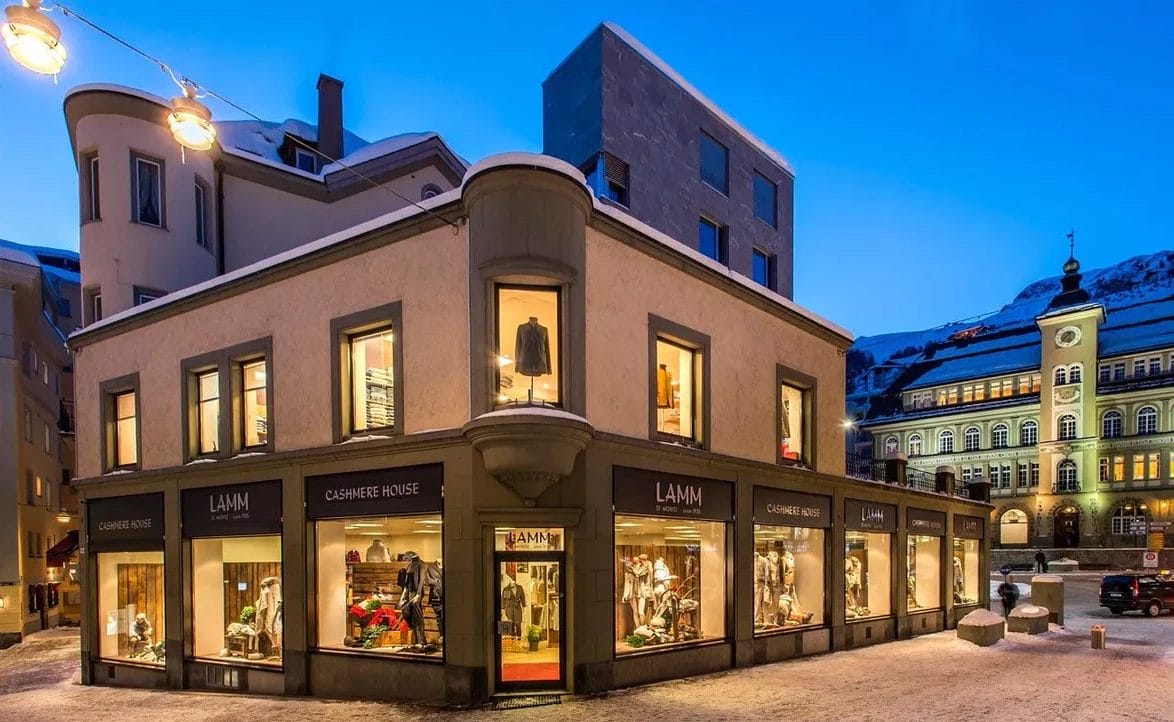 Street view of Lamm Cashmere House at twilight with illuminated display windows, showcasing fashion items in a snowy Alpine town.