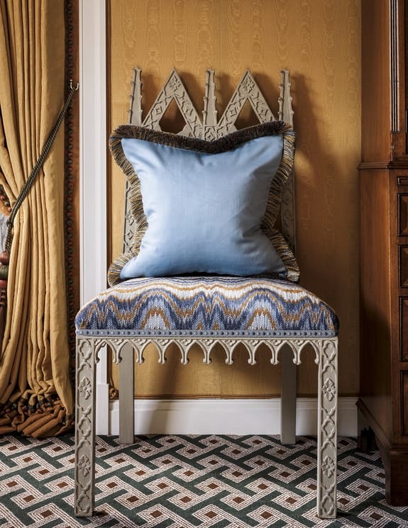 Ornate chair with blue cushion and patterned seat against a patterned carpet and yellow wall with draped curtain.