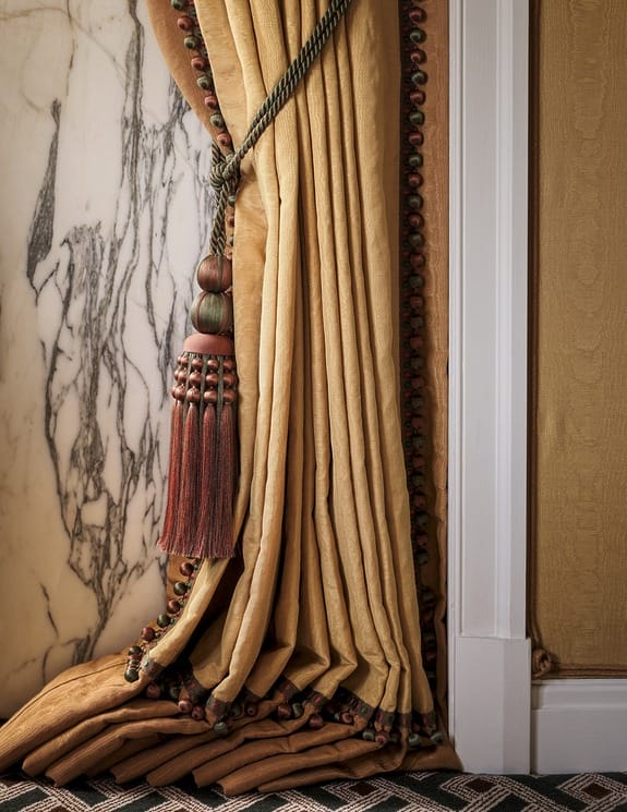 Elegant draped curtain with decorative tassel tieback near marble wall and patterned carpet, creating a luxurious interior setting.