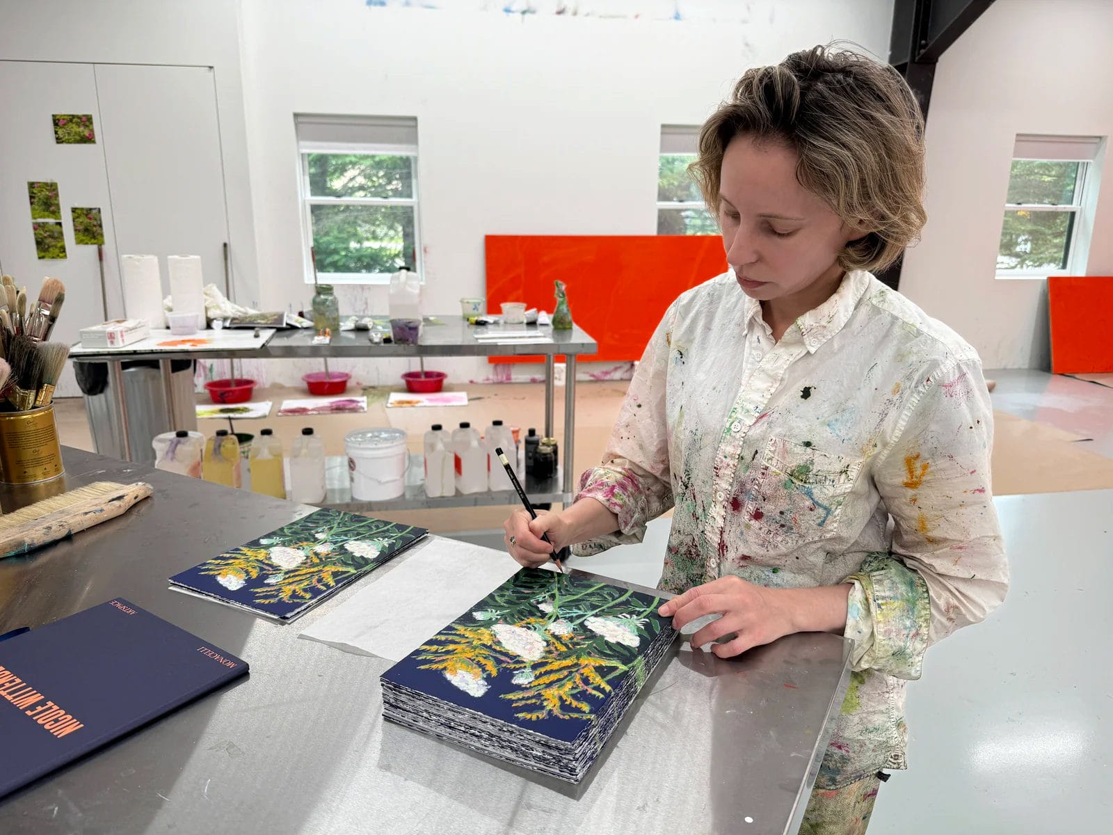 Artist painting botanical prints in a studio filled with art supplies and canvas.
