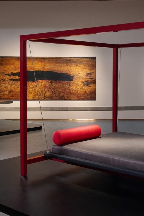 Modern art exhibit featuring a vibrant red-framed bed structure with a cylindrical pillow and a textured painting in the background.