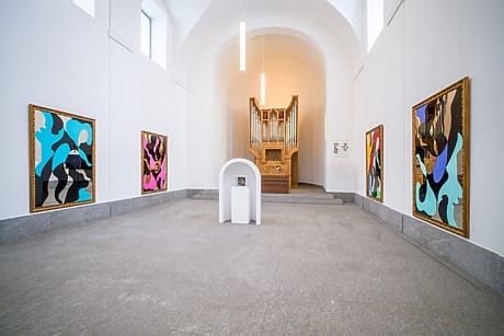 Contemporary art gallery with colorful abstract paintings on white walls and a pipe organ in the background.