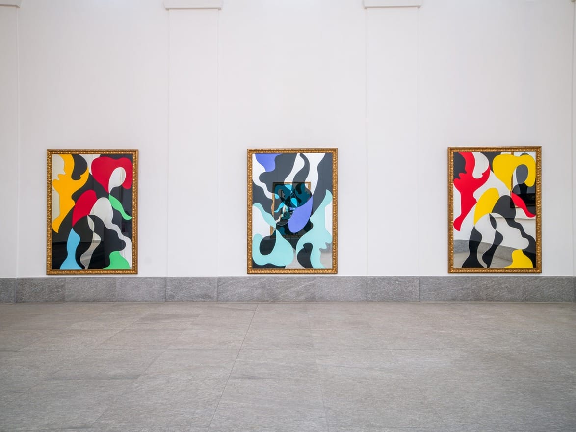 Abstract paintings with vibrant colors in gold frames displayed on a white gallery wall with a gray tile floor.