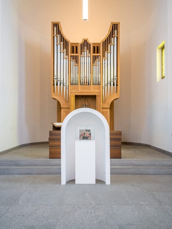 Contemporary church interior with a modern pipe organ behind a small white pedestal displaying artwork on a gray floor.