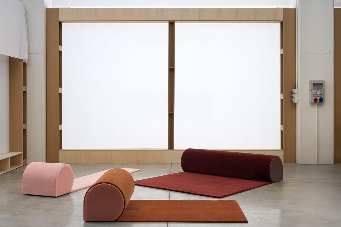 Minimalist room with various colored cylindrical cushions on a concrete floor, and a large backlit panel in the background.