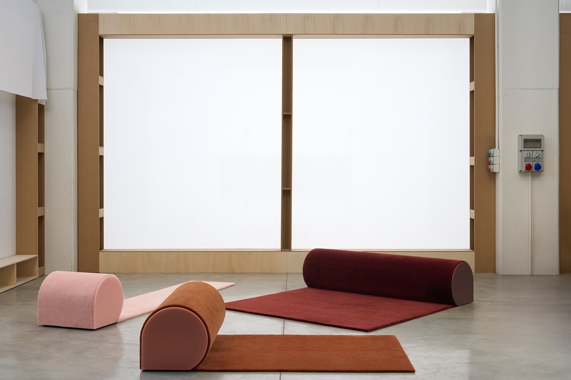 Minimalist room with various colored cylindrical cushions on a concrete floor, and a large backlit panel in the background.