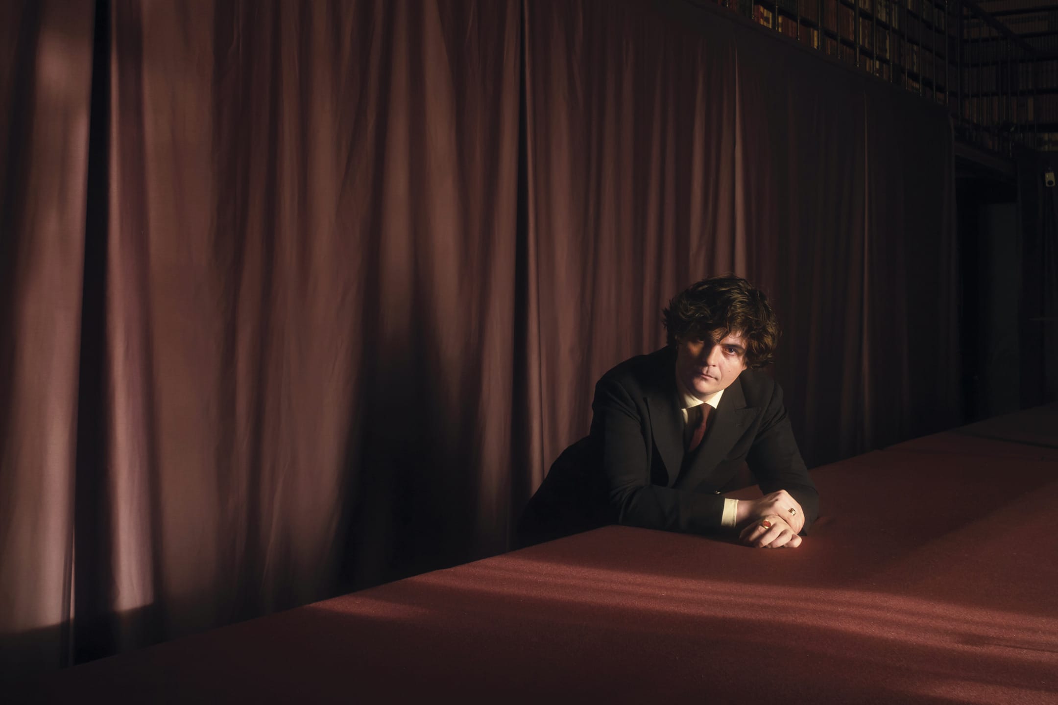 Man in a suit sits alone at a long table with a soft maroon curtain backdrop, exuding a contemplative mood.