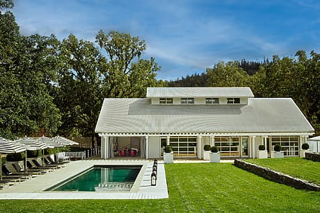 Modern house with a white facade, large windows, pool, lounge chairs, and umbrellas, surrounded by lush greenery and trees.