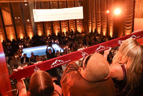 People at an indoor event toasting with a long wooden board, with a pool and a crowd in the background.