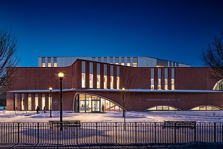 Modern building exterior at night with illuminated windows and snow on the ground, surrounded by bare trees and lamp post.