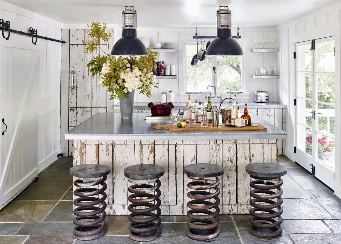 Rustic kitchen with metal bar stools, weathered wood island, hanging lights, and floral arrangement.