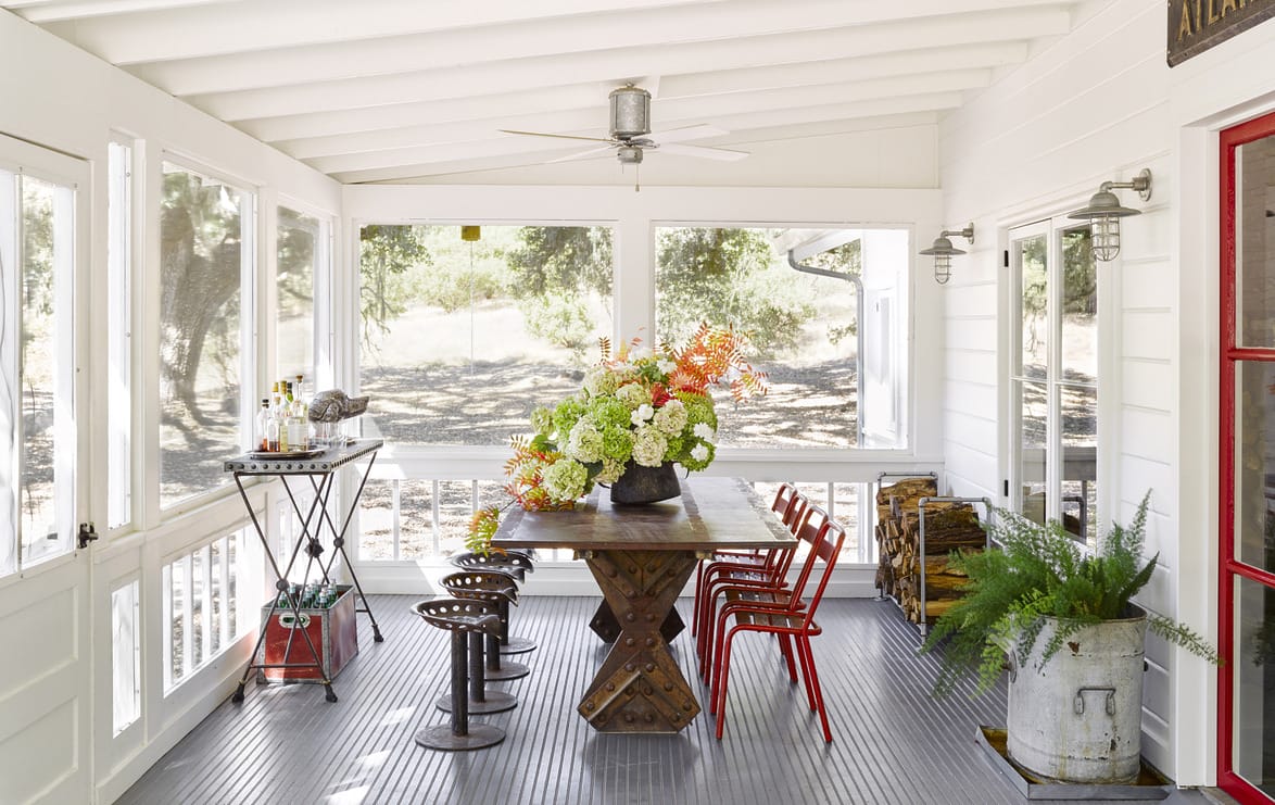 Bright screened porch with wooden table, red chairs, and vibrant floral centerpiece, surrounded by large windows and natural light.