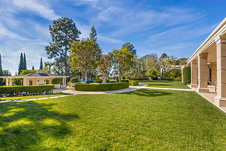 Spacious backyard with manicured lawn, trees, shrubs, and a small garden structure, under a clear blue sky.
