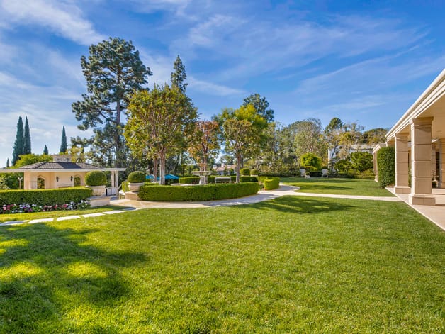 Spacious backyard with manicured lawn, trees, shrubs, and a small garden structure, under a clear blue sky.