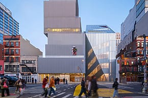 People crossing the street in front of a modern, multi-level building at dusk in an urban cityscape.