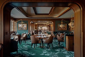 Elegant restaurant interior with wooden accents, green carpet, and leather chairs arranged around set dining tables.