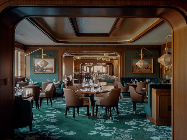 Elegant restaurant interior with wooden accents, green carpet, and leather chairs arranged around set dining tables.