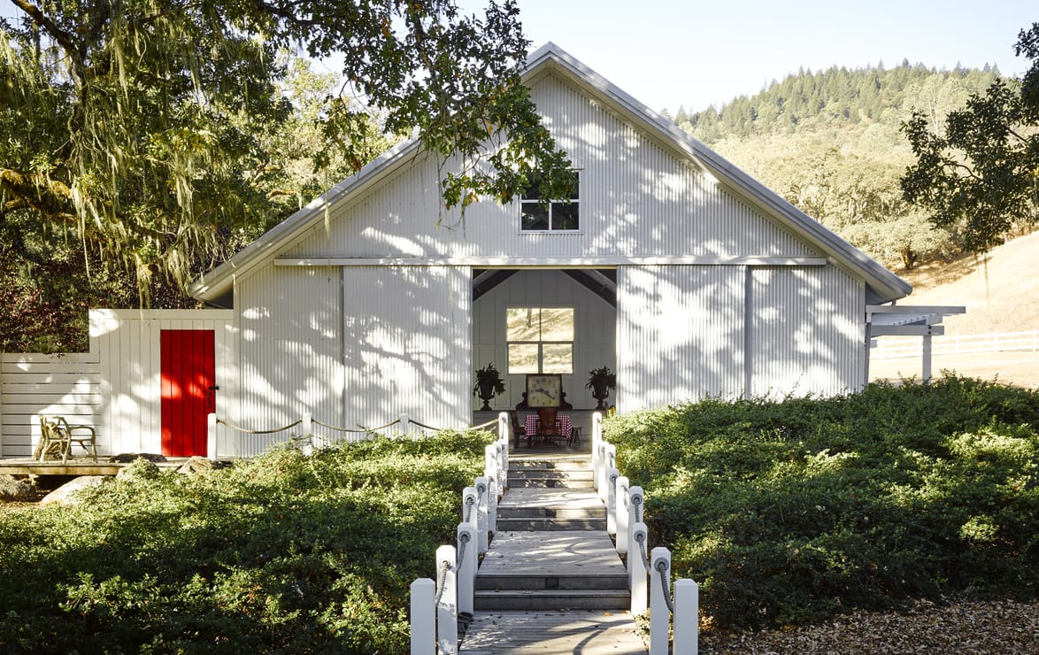 A white barn with a bright red door surrounded by greenery and trees, with a clear blue sky in the background.