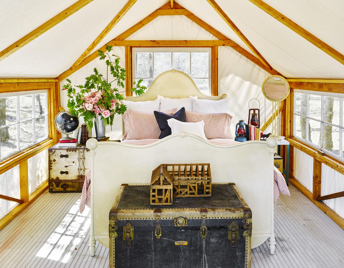 Rustic bedroom with vintage decor, white linens, wooden bedframe, floral arrangement, globe, lantern, and large trunk.