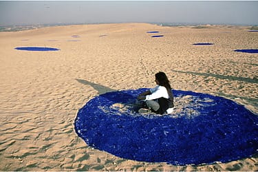Person sitting on large blue circle in sandy desert with several smaller blue circles scattered around.