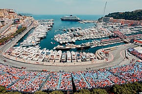 Aerial view of a busy marina with numerous yachts and a racetrack filled with spectators, set against a coastal city backdrop.