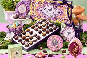 Assorted luxury chocolates in decorative packaging displayed with moss, vibrant Easter eggs, and a purple rabbit ornament.