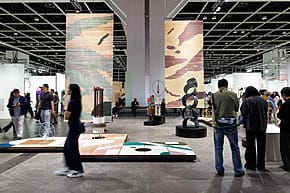 People walking through a modern art exhibition with sculptures and large patterned panels under industrial lighting.