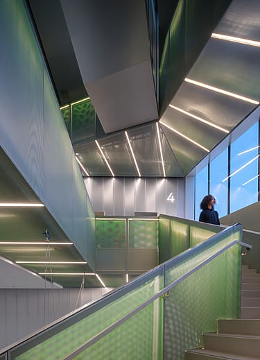 Futuristic staircase with green-lit walls and a person looking upward, surrounded by modern architectural design.
