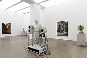 Modern art exhibit with classical statue wired to speakers, surrounded by three large framed photographs on gallery walls.