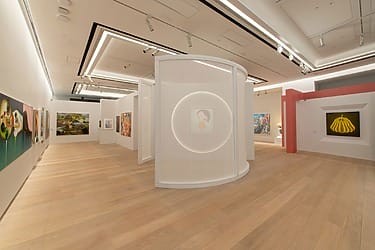 Modern art gallery interior with wooden floors, curved white walls, and various colorful paintings displayed on the walls.