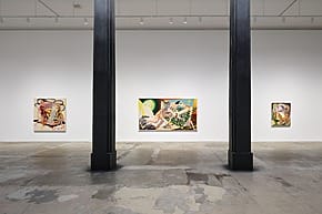 Art gallery interior with abstract paintings on white walls, concrete floor, and two black columns in the foreground.