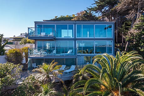 Modern glass house surrounded by lush greenery with ocean view and spacious balcony.