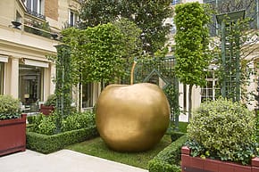 Large golden apple sculpture in a landscaped garden surrounded by greenery and neighboring building facade.